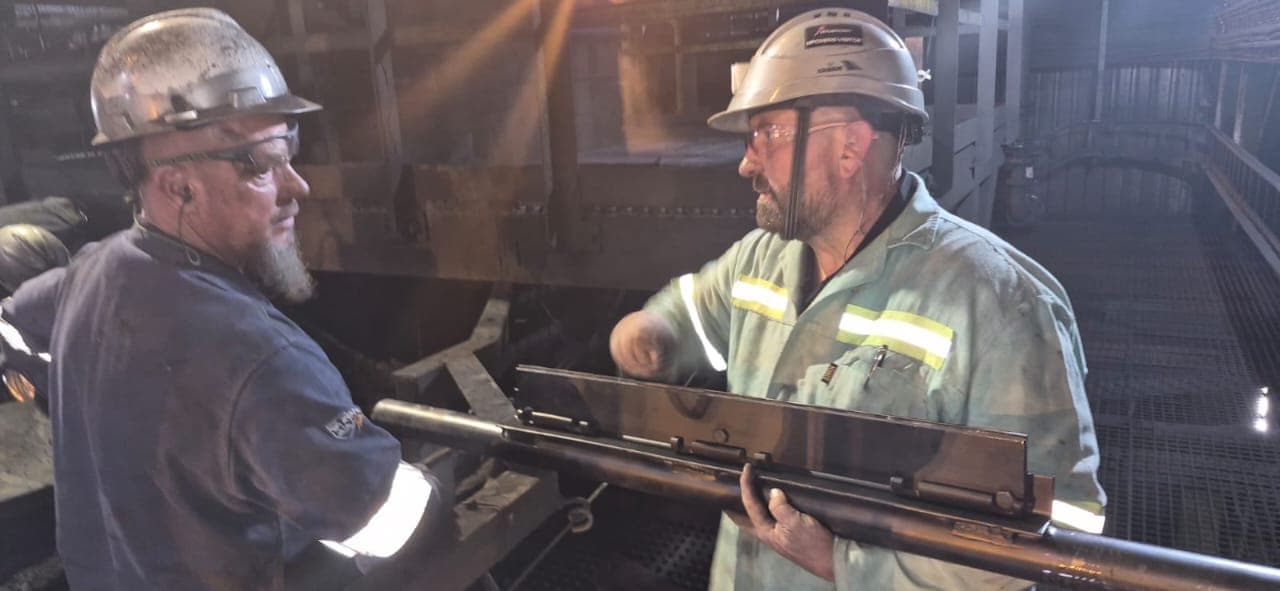Two Kia-Kaha Mining technicians handling a conveyor belt cleaner blade at a transfer point during a site visit